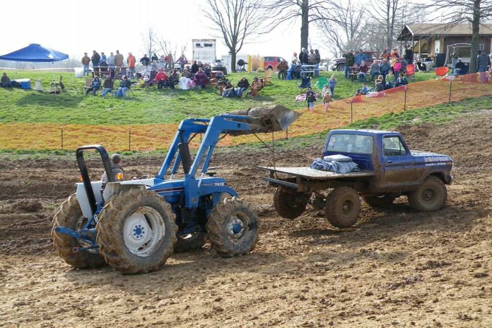 Blue tractor with loader in mud, blue flatbed truck nearby. Spectators watch from grassy hill