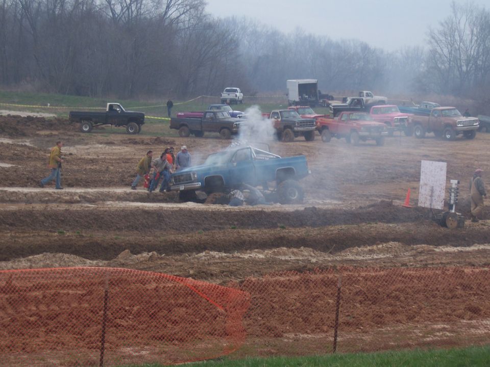 Blue pickup truck stuck in mud, emitting smoke, with people around it