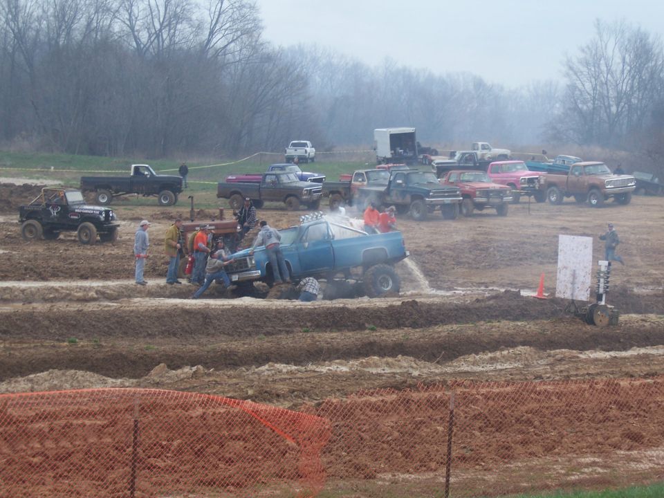 Blue pickup truck stuck in mud pit with people attempting to free it