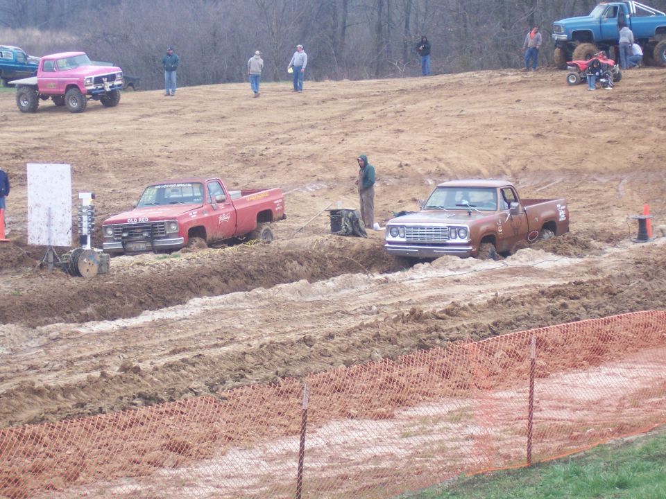 Brown pickup truck stuck in mud, front view. Another red truck is also stuck