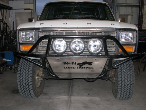 1980 Ford Bronco - The front view of a white Ford Bronco with an aftermarket bumper and four off-road lights is displayed in a garage setting. The bumper has…