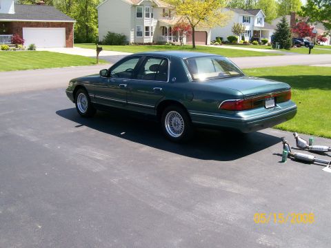 1995 Mercury Grand Marquis - Rear three-quarter view of a dark green 1990s Mercury Grand Marquis four-door sedan parked on a suburban driveway, wearing lace-spoke wire-…