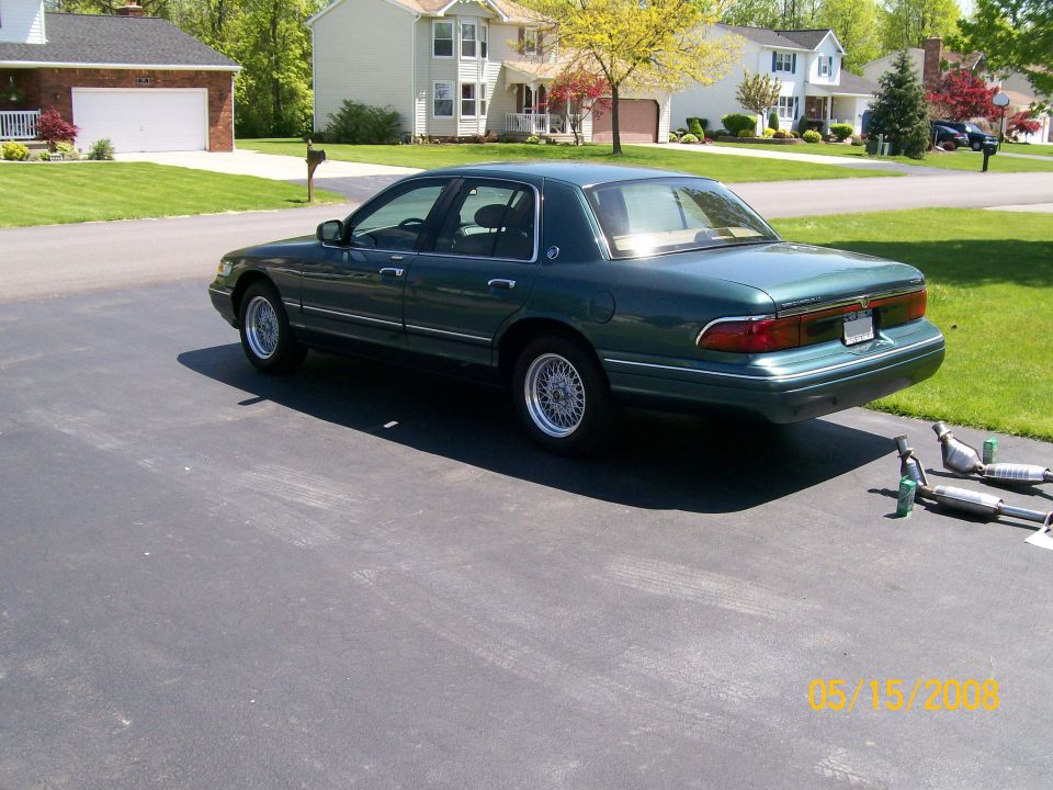 Dark green Mercury Grand Marquis sedan on driveway with removed exhaust parts on the pavement