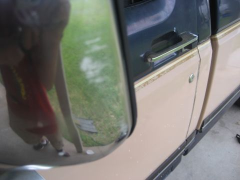 1981 Ford Bronco - A close-up shot of the driver's side door handle and lock on a tan and dark blue pickup truck. The reflection in the side mirror shows a pe…