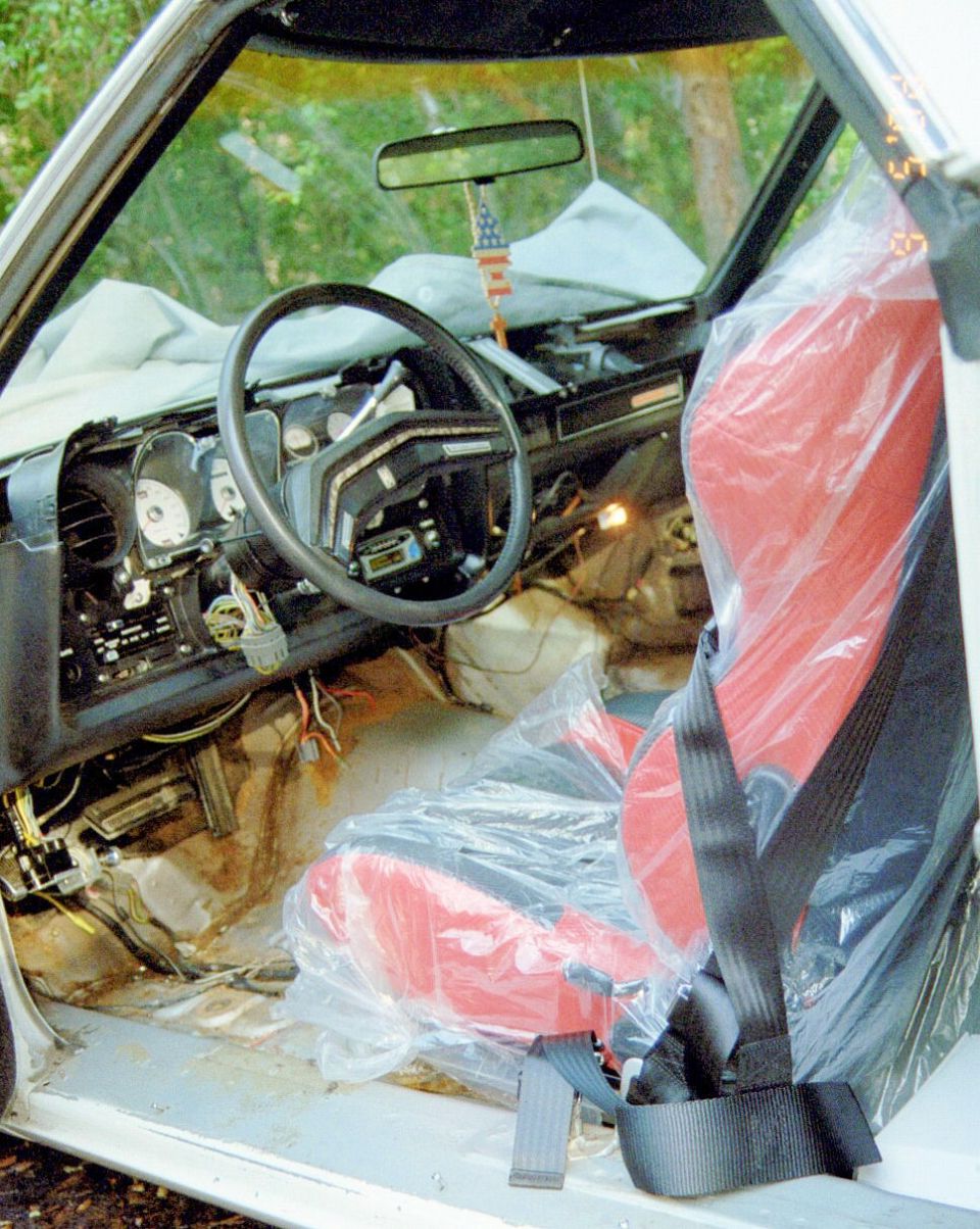 Stripped white car interior with plastic-wrapped red bucket seat, bare floor and exposed gauges