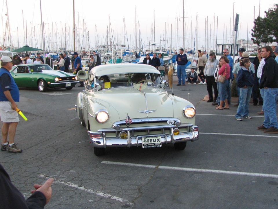 Vintage light-colored sedan with chrome details parked near a marina with people around.