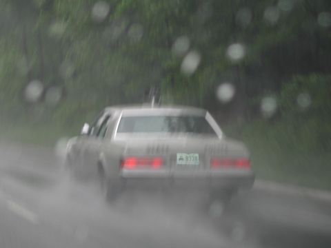 1991 Ford LTD Crown Victoria - tan four-door sedan drives away on a rain-slicked road, photographed through a water-spotted windshield.