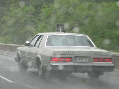 1991 Ford LTD Crown Victoria - beige sedan lettered STATE POLICE with a single roof beacon travels a wet road in the rain.