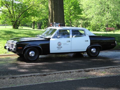 1991 Ford LTD Crown Victoria - black-and-white vintage POLICE sedan with a roof siren parked on a shaded park road by a bench.
