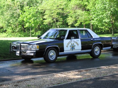 1991 Ford LTD Crown Victoria - black-and-white HIGHWAY PATROL sedan with a star door emblem and push bumper parked on a tree-lined road.