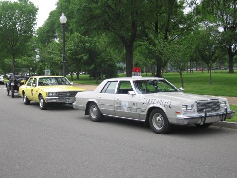 1991 Ford LTD Crown Victoria - silver STATE POLICE sedan with roof beacon parked beside a yellow police car on a tree-lined park road.