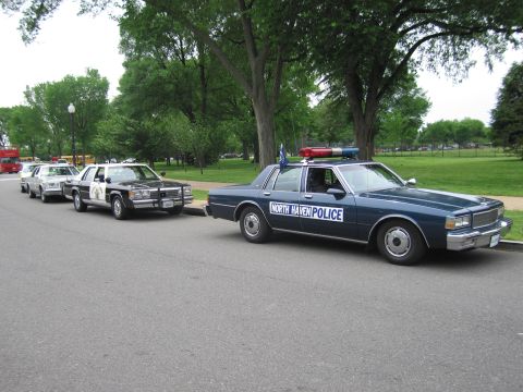 1991 Ford LTD Crown Victoria - dark blue 1980s Chevrolet Caprice marked North Haven Police leads a row of vintage police cars parked along a tree-lined park road.