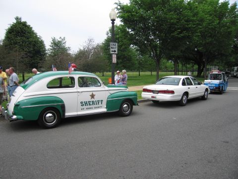 1991 Ford LTD Crown Victoria - green and white vintage Sheriff sedan marked St. Johns County sits in a line of police cars on a park street with onlookers nearby.