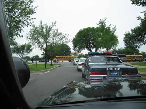 1991 Ford LTD Crown Victoria - View through a windshield of a dark Chevrolet Caprice police car ahead on a road, with yellow school buses parked in the background.