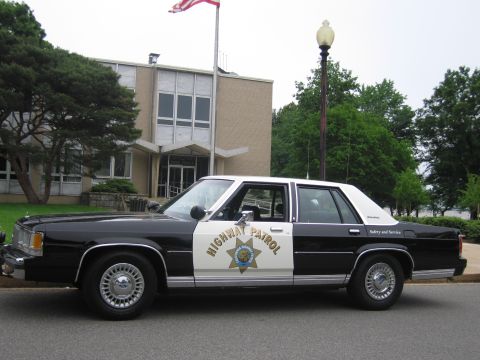 1991 Ford LTD Crown Victoria - black and white Highway Patrol Ford sedan with a gold star door emblem is parked outside a building with an American flag.