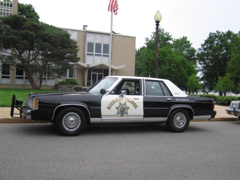 1991 Ford LTD Crown Victoria - black and white Highway Patrol Ford sedan with a front push bar is parked along a curb in front of a low brick building and flagpole.
