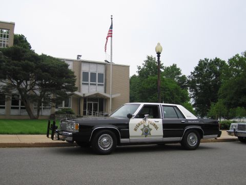 1991 Ford LTD Crown Victoria - black and white Highway Patrol Ford sedan sits beside a flagpole flying an American flag, in front of a brick municipal building.