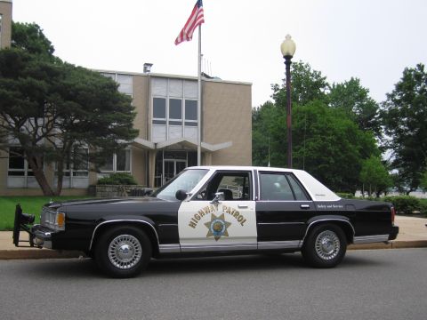 1991 Ford LTD Crown Victoria - Side profile of a black and white Highway Patrol Ford sedan with a chrome push bar, parked outside a brick building with an American flag.