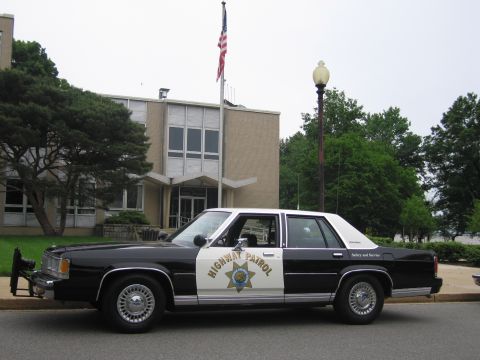 1991 Ford LTD Crown Victoria - black and white Highway Patrol Ford sedan with a front push bar is parked at a curb before a brick building and flagpole.