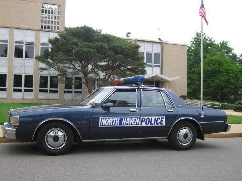 1991 Ford LTD Crown Victoria - dark blue Chevrolet Caprice marked North Haven Police is parked in profile in front of a brick building, flagpole, and pine tree.