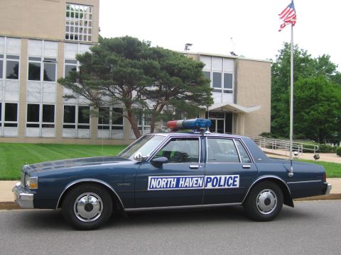 1991 Ford LTD Crown Victoria - dark blue Chevrolet Caprice marked North Haven Police, topped with a lightbar, sits before a brick building with an American flag.