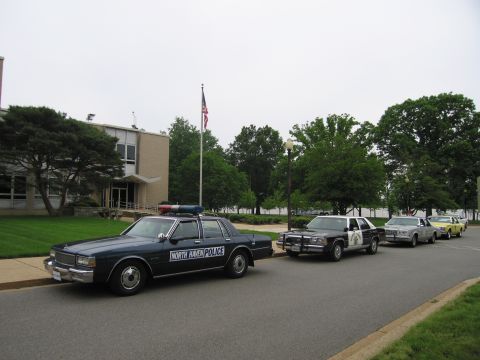 1991 Ford LTD Crown Victoria - dark blue North Haven Police Caprice leads a line of vintage police cars parked along a curb before a brick building and flagpole.