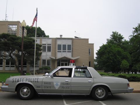 1991 Ford LTD Crown Victoria - silver sedan marked Rhode Island State Police, topped with a red light, is parked outside a multi-story stone building with a flag.