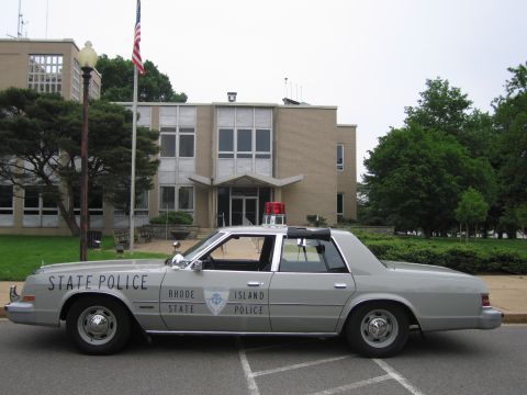 1991 Ford LTD Crown Victoria - silver Rhode Island State Police sedan with a single red roof light sits in profile before a tan brick building and flagpole.