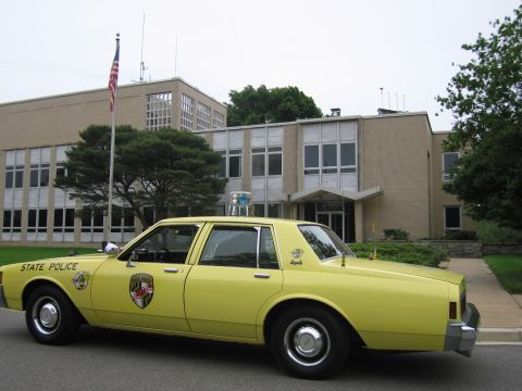 1991 Ford LTD Crown Victoria - bright yellow Chevrolet Impala sedan marked State Police is parked in profile before a tan brick municipal building and flagpole.