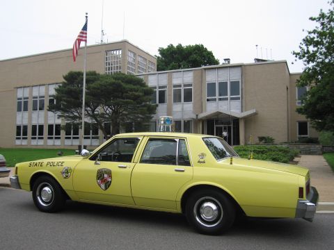 1991 Ford LTD Crown Victoria - yellow State Police Chevrolet sedan with a circular door emblem is parked outside a tan brick building flying an American flag.