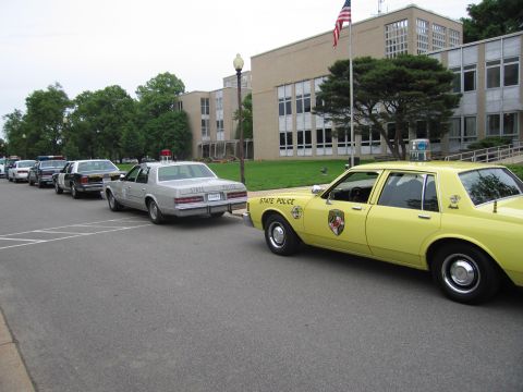1991 Ford LTD Crown Victoria - yellow State Police sedan parks beside a silver sedan in a curbside row of cars before a large brick municipal building.