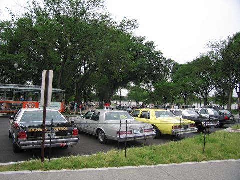 1991 Ford LTD Crown Victoria - lineup of classic police cars, including a black Ford marked Highway Patrol, parked along a tree-lined park drive.