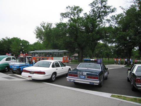 1991 Ford LTD Crown Victoria - white Ford police sedan and a dark blue sedan with 'EMER 911' plate sit near an old trolley in a park parking lot.