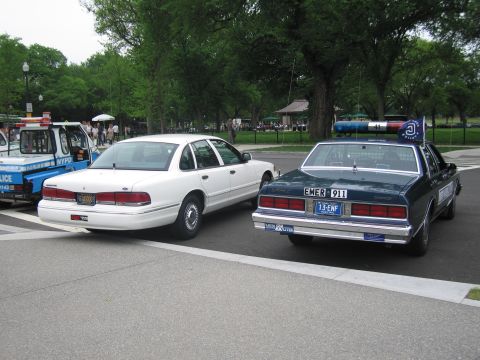 1991 Ford LTD Crown Victoria - Rear view of a white Ford police sedan and a dark blue sedan bearing an 'EMER 911' license plate in a park.