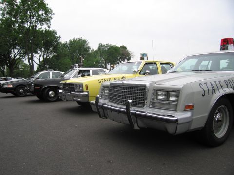 1991 Ford LTD Crown Victoria - row of vintage State Police sedans, including yellow and silver cars with roof beacons, parked at angle in a lot.