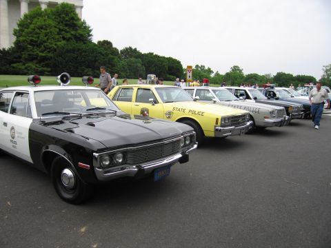 1991 Ford LTD Crown Victoria - diagonal lineup of vintage police cars, black-and-white and yellow State Police sedans, with people standing nearby.