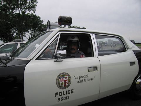 1991 Ford LTD Crown Victoria - mustached man in a white motorcycle-style helmet sits in a white LAPD car, door reading 'to protect and to serve' POLICE 85012.