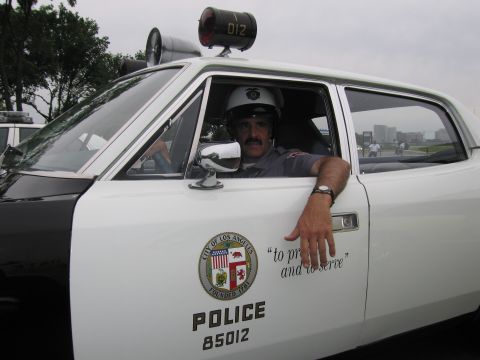 1991 Ford LTD Crown Victoria - mustached man in a white helmet rests his arm out the door of a white LAPD car marked POLICE 85012 with roof speaker 012.