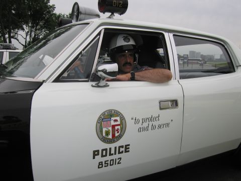 1991 Ford LTD Crown Victoria - man in a white helmet sits in a white LAPD police car, its door bearing the city seal and 'to protect and to serve'.