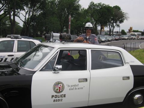1991 Ford LTD Crown Victoria - mustached officer in a white helmet stands beside a white LAPD police car marked POLICE 85012 with a roof loudspeaker.