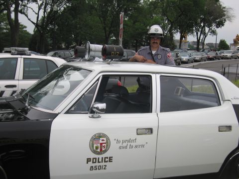 1991 Ford LTD Crown Victoria - officer in a white helmet stands at the open door of a white LAPD police car reading 'to protect and to serve' POLICE 85012.