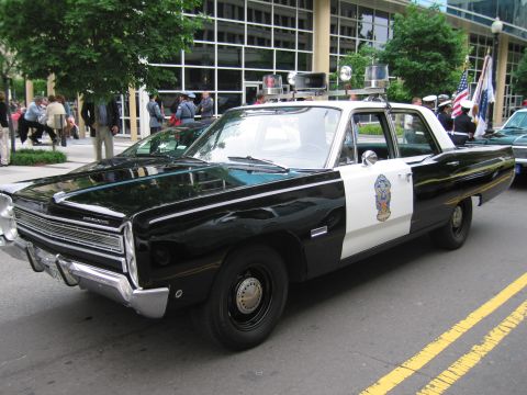 1991 Ford LTD Crown Victoria - black-and-white Plymouth Fury police sedan with multiple roof beacons parked along a city street near onlookers.