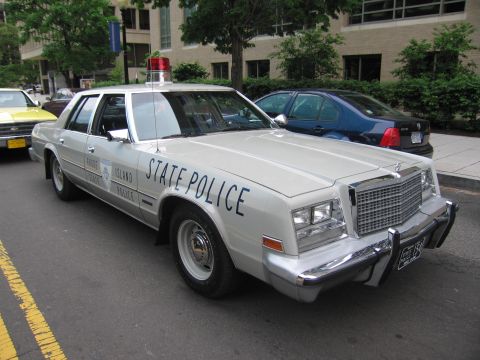 1991 Ford LTD Crown Victoria - cream-colored Chrysler sedan lettered 'STATE POLICE' with a red roof beacon parked along a downtown street.