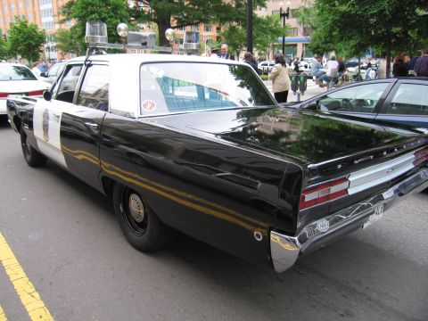 1991 Ford LTD Crown Victoria - Rear quarter view of a black Pontiac police sedan with a gold body stripe and roof beacons parked along a city street.