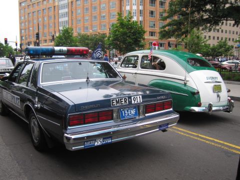 1991 Ford LTD Crown Victoria - dark blue Chevrolet Caprice police car marked 'EMER 911' parked beside a green vintage 'Sheriff' woody-style car downtown.