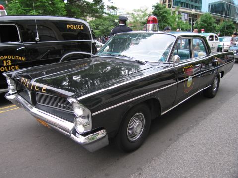 1991 Ford LTD Crown Victoria - black 1960s Pontiac police sedan lettered POLICE with a roof beacon, an officer in cap standing behind it on a city street.
