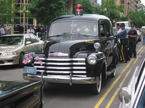 1991 Ford LTD Crown Victoria - restored black 1950 Chevrolet police car marked POLICE sits on a city street with small American flags and uniformed officers beside it.