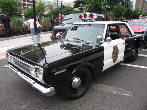 1991 Ford LTD Crown Victoria - black-and-white mid-1960s Plymouth police cruiser with twin red roof beacons and a star door emblem sits at a downtown car show.