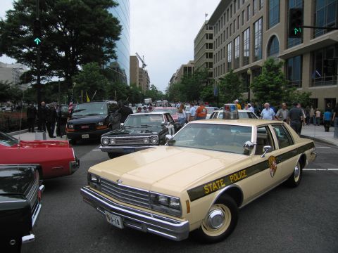 1991 Ford LTD Crown Victoria - tan late-1970s State Police four-door sedan with a brown stripe and gold door shield is parked among other classic cars downtown.