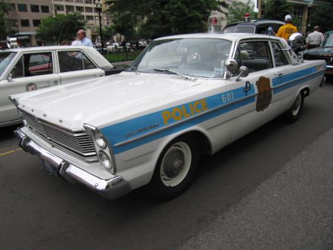 1991 Ford LTD Crown Victoria - white mid-1960s police sedan with a blue stripe lettered POLICE 601 and a door shield is displayed on a city street.